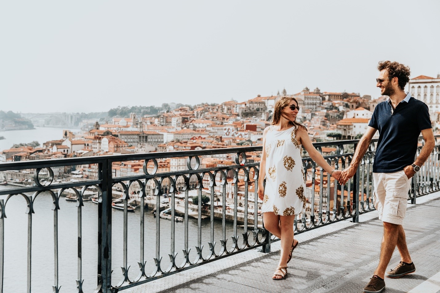 Man and woman walking on bridge with port city behind them