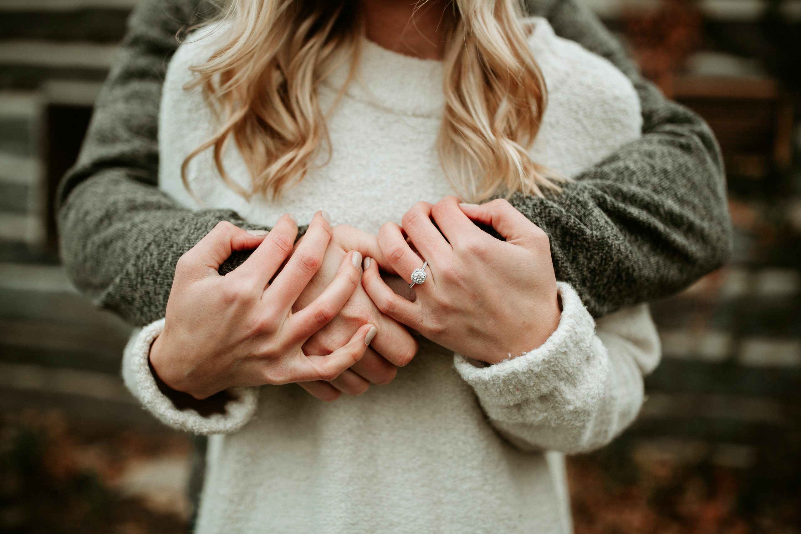 A person in a light sweater embraces another person from behind, their hands interlocked with a visible ring on one hand.